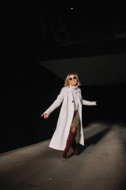 Güneş gözlüklü ve uzun paltolu bir kadın gölgeli bir şehir koridorunda neşeyle yürüyor, kendine güveni tam.