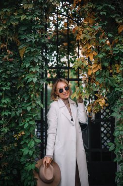 Şık paltolu bir kadın, elinde şapkasıyla canlı yeşilliklerle dolu bir kapının yanında duruyor..