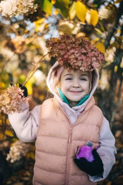 Neşeli bir çocuk parkta oynuyor, elinde renkli yapraklarla bir dal tutarken sonbahar güzelliği ile çevrili..