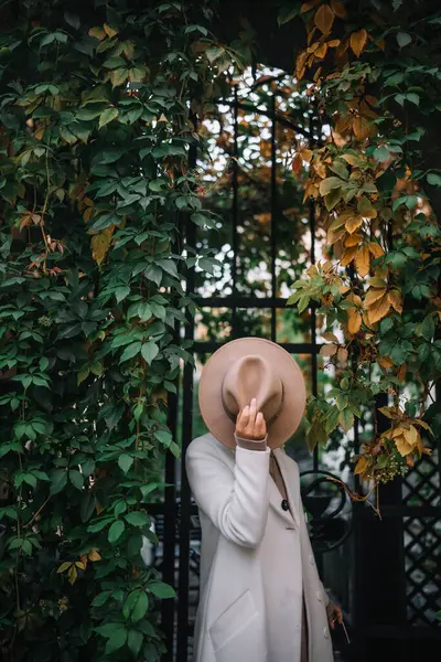 Beyaz önlüklü ve şapkalı bir kadın, canlı sonbahar yapraklarıyla çevrili bir kapının önünde neşeli bir şekilde poz veriyor..