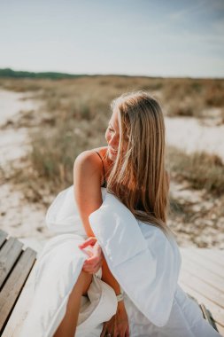 Sandy Area 'daki tahta güvertede beyaz battaniyeye sarılı uzun sarı saçlı genç bir kadın oturuyor. Sakin yaz tatili konsepti