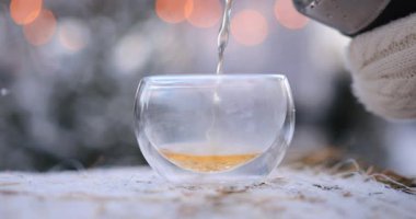 Kadın elinde sıcak beyaz eldivenlerle termostan dışarıya dobble duvarlı cam bardağa siyah çay döküyor. Sıcak içecekle kış pikniği, güzel çelenkler ve arka planda yağan kar.