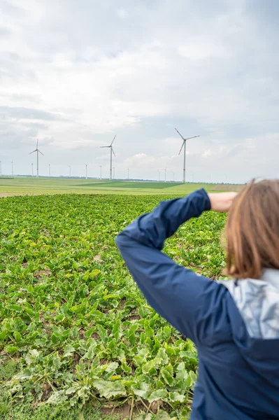 Woman with brown hair and blue jacket is looking at a wind park from the distance, wind park with wind turbines in agricultural field area, rear view, vertical shot