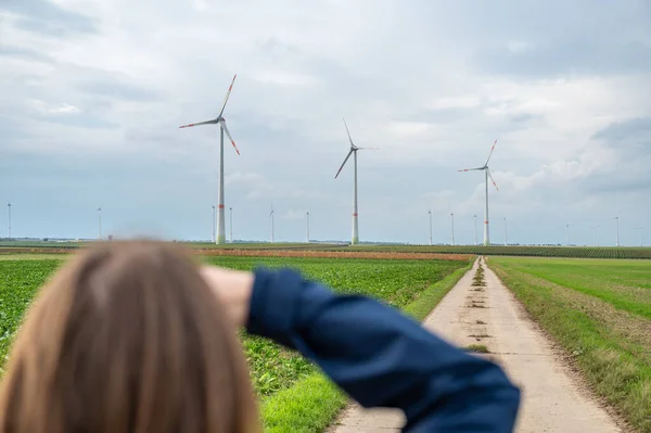 Woman with brown hair and blue jacket is looking at a wind park from the distance, agricultural path in front wind turbines in agricultural field area, rear view, vertical shot