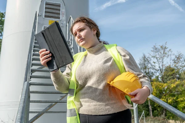 Ingeneer boss woman with yellow helmet and brown curly hair is inspecting a wind turbine with her digital tablet, worried facial expression