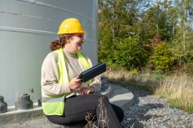 Female engineer with brown curly hair and yellow helmet is smiling and laughing while sitting on the ground of a wind turbine with the screws in background, holding a digital tablet