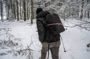 Man with hat and backpack is hiking trough the forest during winter with lots of snow, rear view