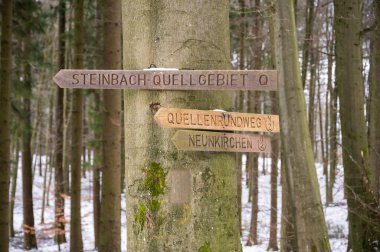 Wooden way sign path sign Steinbach-Quellgebiet, Quellenrundweg and Neunkirchen hanging on a tree during winter with snow in background