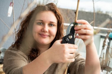 Woman with brown curly hair is cutting the fastening strap between plant and plant support with a secateurs, smiling and looking at camera, sitting on the balcony