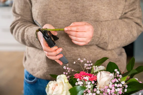 Female florist with jeans and beige pullover is cutting a plant with a secateurs indoors, beautiful bunch of flowers in the front, no visible face