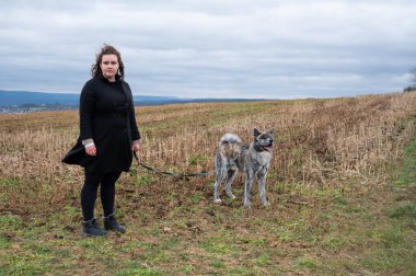 Kahverengi kıvırcık saçlı ve siyah ceketli kadın bulutlu bir günde kırsalda gri kürklü köpeğini gezdirirken kameraya bakıyor.