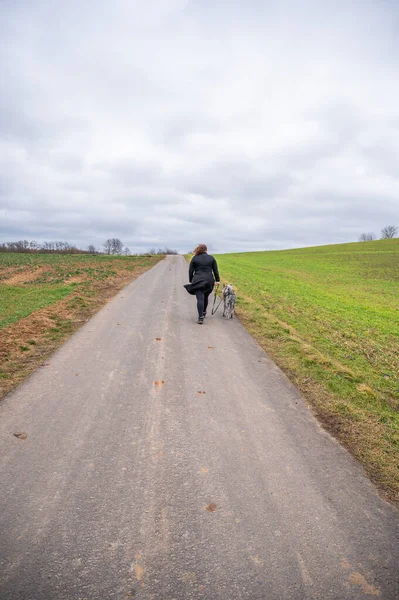 Woman with brown curly hair and black jacket walking her akita inu dog with gray fur on an agricultural path during cloudy day, vertical shot, view from the distance