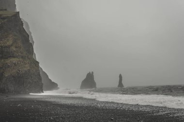 Kara kumsal Reynisfjara, İzlanda sisli havada, önünde deniz ve sahil olan dağ sıraları
