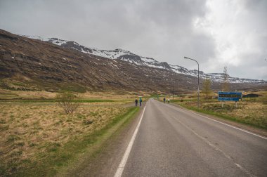 Caddenin yanında yürüyüş yapan ve Seydisfjordur yönünde yürüyen turistler, arka planda kar dağı sıraları, İzlanda