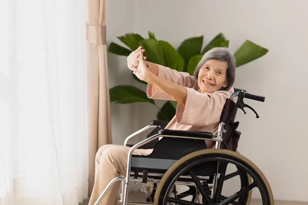 高齢のアジア系女性は自宅のリビングで車椅子で運動をしています — ストック写真