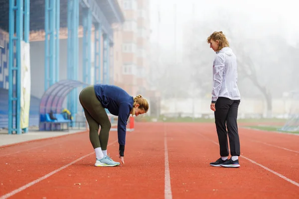 霧深い朝 2人の若い女性がコース上でストレッチの足を走らせている 陸上競技トレーニングのコーチと訓練生 — ストック写真