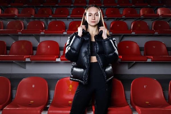 Young Attractive Sportswoman Wearing Puffer Jacket Standing Tiers Red Seats lizenzfreie Stockfotos