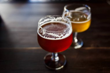 Two glasses of craft beer are placed on a wooden table in a cozy bar. One glass contains amber ale, while the other showcases a light lager clipart