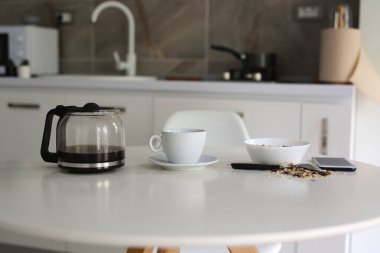 morning in a cozy apartment, on a white table a kettle with fresh black coffee, a white cup and muesli in a plate, a mobile phone lies nearby
