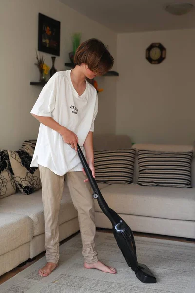 13歳の10代の男が家を掃除し 床を掃除し ソファを掃除し 少年は家を掃除する 青いミトンとベージュの服を着た男子供が窓を洗ってほこりを拭く — ストック写真