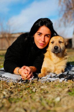 Siyah kazaklı, güzel, koyu renk saçlı bir kız bir köpekle, evsiz bir köpekle, bahçede çimlerin üzerinde uzanmış, sarılıyor, telefonda fotoğraf çekiyor. Kız evsiz bir köpeği umursuyor.