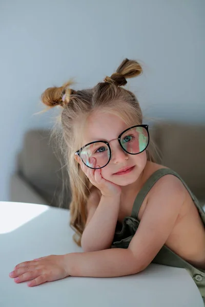 一个漂亮的 时髦的三岁小女孩 戴着眼镜 坐在一张白色的桌子旁 想象着 看着相机 摆出一副摄影师的样子 — 图库照片