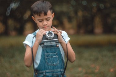 Genç bir fotoğrafçı: Fotoğraf sanatından büyülenen genç bir çocuk, elinde antika bir kamera tutuyor, gözleri objektife odaklanmış, bir anı yakalamaya hazır. Sahne yumuşak, doğal bir banyo.