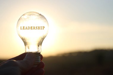 Hand holding light bulb with the text leadership in front of the bright sun clipart