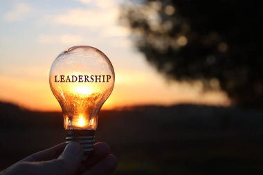 Hand holding light bulb with the text leadership in front of the bright sun clipart