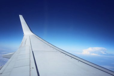 Mavi gökyüzüne ve bulutlara karşı pencereden bir uçağın kanat görüntüsü.
