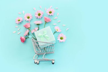 Çiçekli alışveriş arabası ve pastel mavi arka planda hediye kutusu. Tatil alışverişi ve satış konsepti
