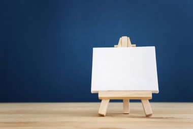 Bir standın üzerinde küçük bir tuval, mavi arka planlı ahşap bir masanın üzerine yerleştirilmiş.