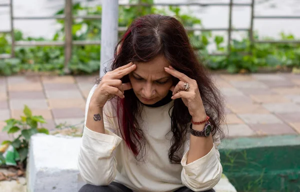 Depressed worried Woman sitting in stress pose in a park bench and looking head down posture. Front view. Indian Ethnicity Age 50 to 54 Years.