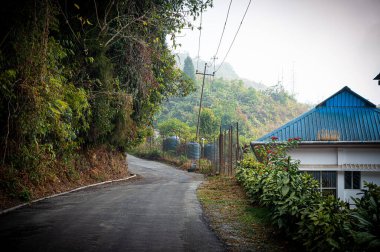 A house near a hill side road going uphill on a mountain valley against mountain forest and clear sky in the background.