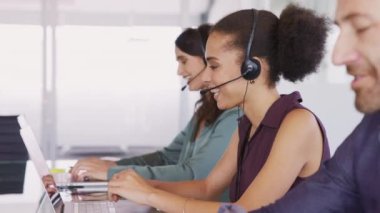 Smiling multiethnic people with headsets using computer and smiling while working in office. Young man and woman operators talking on headset with clients. Group of telemarketing customer service team working while sitting in a row.