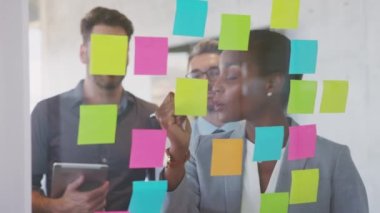 Smiling black business woman writing on sticky notes on glass wall in meeting room. Business people planning and discussing using post it notes in modern office. Black leader showing strategy plan to colleagues during brainstorming.