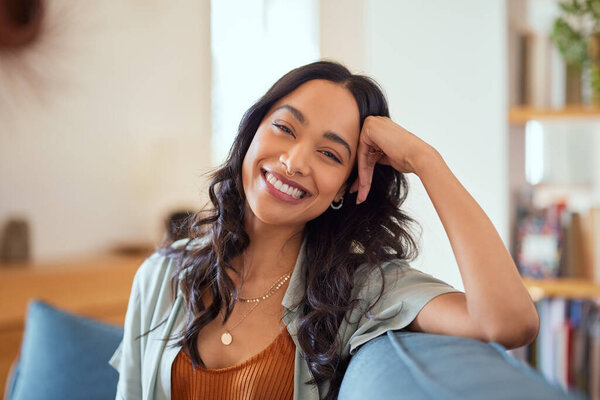 Portrait of happy young latin woman sitting on sofa at home while looking at camera. Happy multiethnic carefree woman in casual clothing relaxing at home sitting on couch. Cheerful mexican girl sitting on couch and laughing.