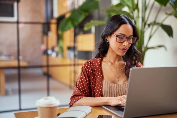 聚精会神的拉丁女商人戴着眼镜在笔记本电脑上工作 在办公室大堂的笔记本电脑上工作的有重点的女企业家 年轻而认真的多族裔女商人戴着眼镜在网上工作 图库图片