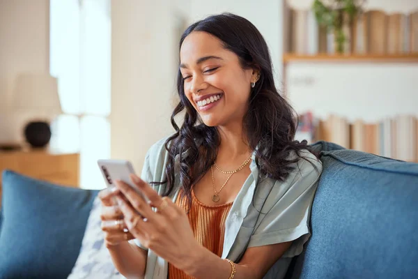 愉快的拉丁女人在家里坐在沙发上的视频通话中使用智能手机 用智能手机在社交网络上与他的恐慌女孩聊天 年轻的混血儿一边笑一边看手机上有趣的视频 图库图片