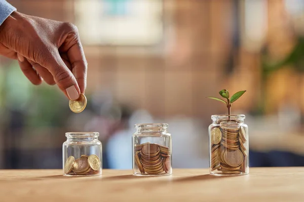 Close up of black woman hand adding money in coin in a jar. Girl hand holding coin adding money in glass jar of different sizes. Investment, savings and interest concept.