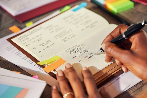 Closeup of african american woman hands making a list of work in diary on desk. Rear close up view of successful woman planning her week in advance while writing in personal organizer, using colored sticky notes. Detail of casual woman listing down t