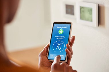 Evdeki akıllı telefonu kullanarak siyah kadın elinin klima ya da ısı pompasının ısısını ayarladığı yeri kapatın. Afrika kökenli Amerikalı bir kadının oturma odasındaki akıllı ev kontrol uygulamasını kullanarak ısıyı ve havalandırmayı ayarlıyor. Uzaktan denetim o