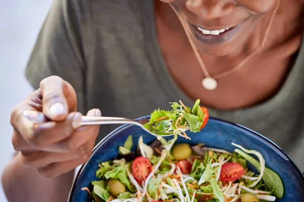 Close up of happy black woman eating healthy salad with green fresh ingredients. Smiling vegetarian woman holds bowl of fresh salad while eating tomatoes and carrot with fork. Detail of beautiful african american lady eating vegan food at home during