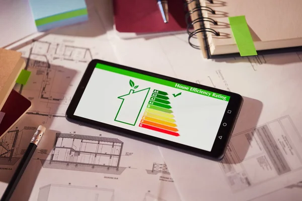 Energy efficiency screen on cellphone leaning on drawings and blueprints of a new house. Architect\'s desk with plans on it and a cell phone with an energy efficiency graph. Renewable energy, renovation, energy class and eco house concept.