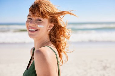 Plajda güneş kremi sürmüş güzel genç bir kadın kameraya bakıyor. Denizde güneş kremi sürerken yaz tatilinin tadını çıkaran kızıl saçlı güzel kız. Sağlıklı ve narin ciltli, yanağında güneş kremiyle fotokopi makinesiyle mutlu bir kadının portresi.