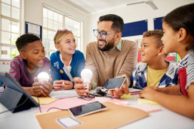 Güneş paneli olan mutlu erkek öğretmen güneş enerjisini aydınlatan ampullerle açıklıyor. Fen bilgisi sınıfındaki öğretmen elektrik üretmek için güneş panelini gösteriyor. Gülümseyen çok ırklı ilkokul çocukları yenilenme konusunda deneyler yapıyorlar.