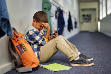 Sırt çantalı küçük bir çocuk akıllı telefonuyla okul koridorunda tek başına oturuyor ve telefonunda aşağılayıcı bir mesaj okuduktan sonra ağlıyor. Üzgün çocuk, ilkokulda yerde tek başına oturmuş, yüzünü eliyle kapatırken hayal kırıklığına uğramış, yalnız hissediyordu., 