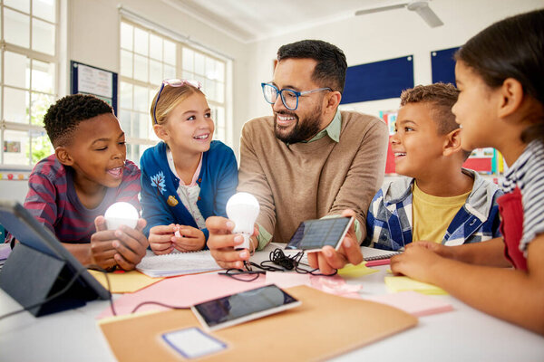 Happy male teacher with solar panel explaining pupil about solar energy with illuminated bulbs. Teacher in science classroom showing a solar panel to generate electricity. Smiling multiethnic primary children learning and experimenting about renewabl