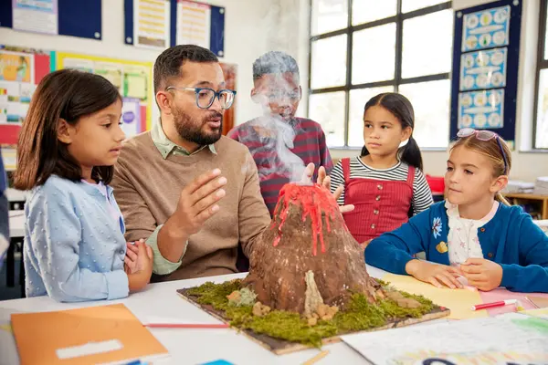 stock image Male teacher teaching to a group of school children about volcanos at science project in school. Group of students understanding volcanic eruption in class with teacher during science class while observing a volcano model that actually erupts with th