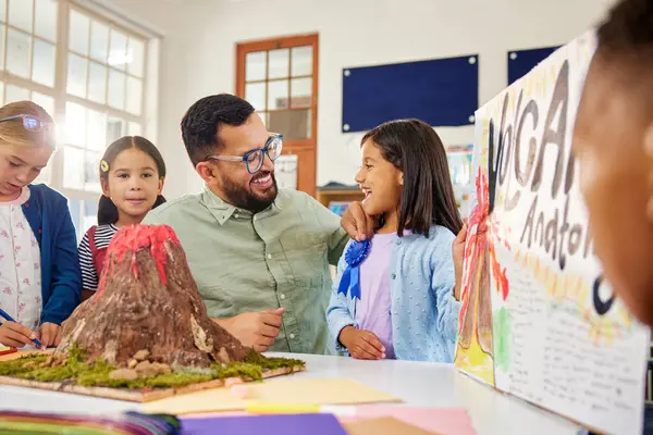 stock image Teacher presenting award badge to indian girl for volcano anatomy explanation in school. Excited little middle eastern girl explaining volcanic eruption using model and chart to her classmates. School girl wins first prize for a science project at pr